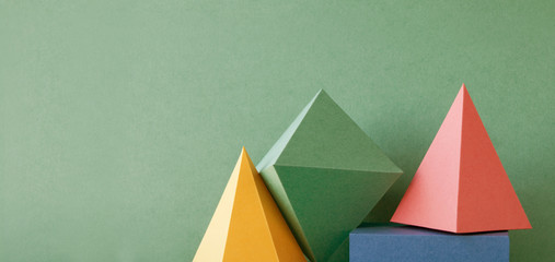 Colorful abstract geometric background with three-dimensional solid figures. Pyramid prism rectangular cube arranged on green paper. Yellow blue pink malachite colored geometrical shapes. Soft focus