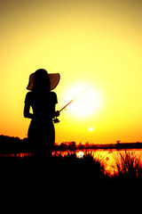 Silhouette of the figure of awoman in a hat with a fishing rod on the river bank