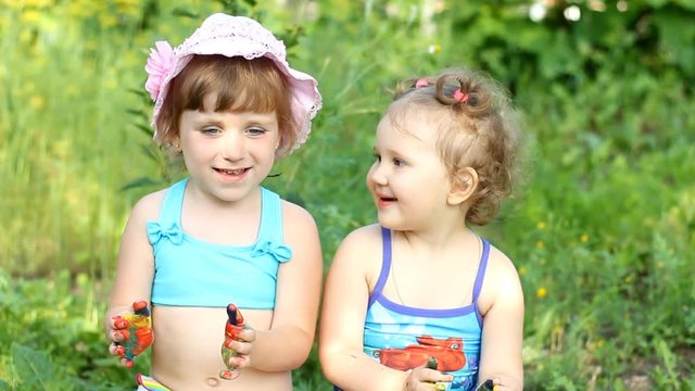 Two happy girl-sisters are sitting in nature in a swimsuit and paint with colors