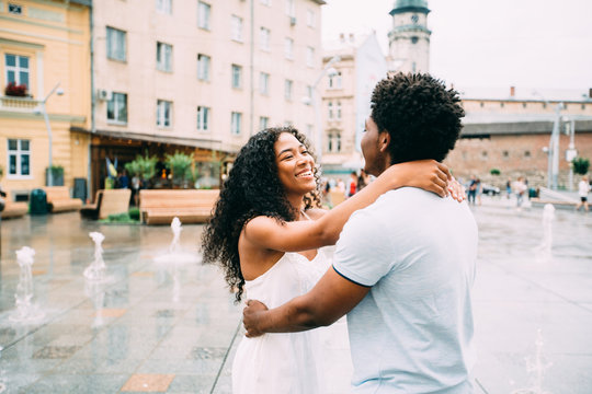 Positive emotional photo of amazing young africam american couple whirling and hugging over city square with fountain on background. Travel recreation lifestyle, water fountain.