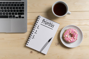 Laptop, pen, coffee, donuts and notebook written with Priorities word on wooden table, business concept.