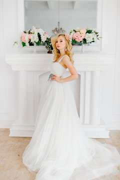 Young beautiful bride looking at camera in white expensive wedding dress in front of decorative fireplace and wall. Preparation for marriage.  Makeup and hairstyle. Happy glamorous blonde angel doll.