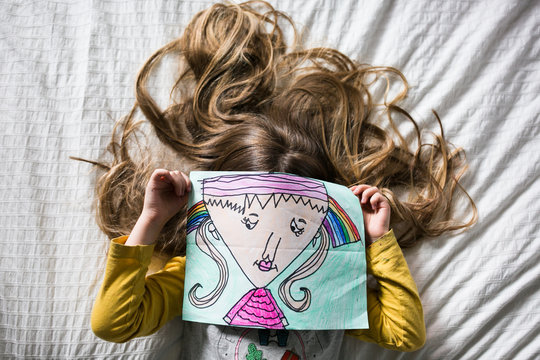Young girl lying on bed, holding drawing of girl in front of face, overhead view