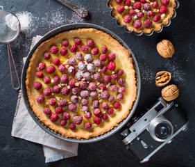 Tart with raspberries, and nuts, powdered sugar on dark stone background with old camera