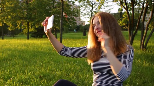 Young woman in striped shirt with long hairs is sitting on grass and taking selfie by using smartphone. Green city park and trees at sunny evening on background.