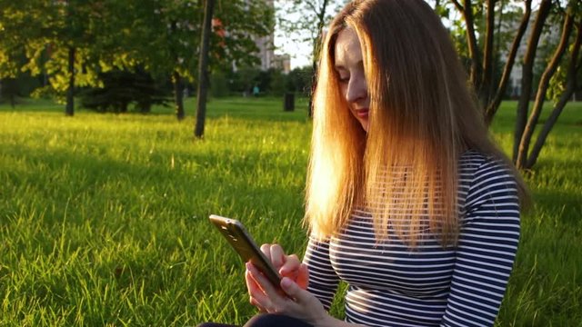 Young woman in striped shirt with long hairs is using smartphone and sitting on grass. Trees and cyclist in green city park at sunny evening on background. Mobile office concept..