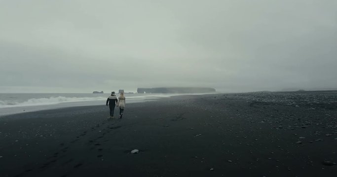 Aerial back view of the young couple in icelandic sweater walking on shore of the sea on black volcanic beach in Iceland