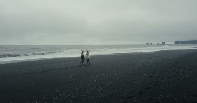 Aerial view of young couple walking, running on black volcanic beach in Iceland. Tourists in lopapeysa enjoy the waves.