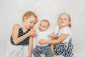 Lifestyle portrait of cute white Caucasian girls sisters holding little baby, lying on bed blanket indoors. Older siblings with younger brother sister newborn. Family love bonding together concept.