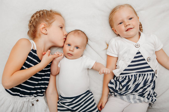 Lifestyle portrait of cute white Caucasian girls sisters holding kissing little baby, lying on bed indoors. Older siblings with younger brother sister newborn. Family love bonding together concept.