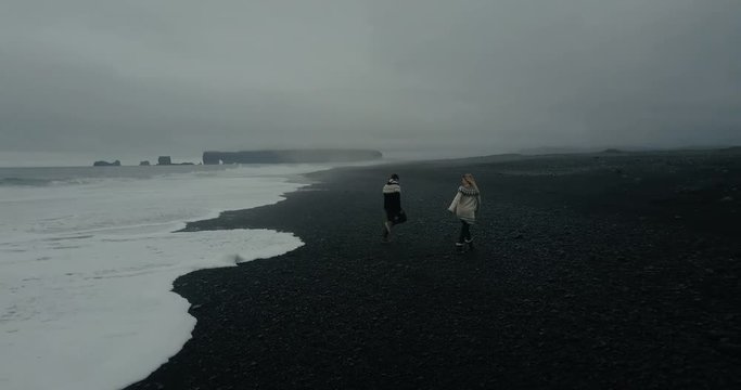 Aerial view to the young couple on the black volcanic beach in Iceland. Man runs from the wave on the shore, woman waits