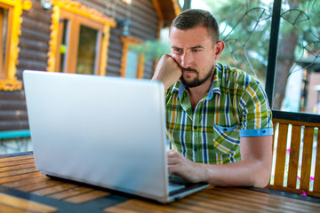 The man works on a laptop. Thoughtfulness and anxiety emotions.On the background of a wooden cottage