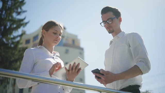 Business partners standing outside using holding phone tablet computer device black white talking young man woman sunny day street working successful confident smart casual clothing calm tapping urban
