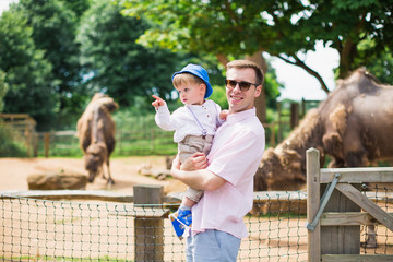 Young man stands in a zoo and keeps his little cute son who looks aside