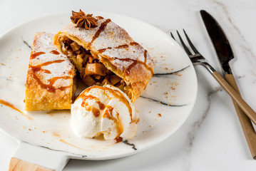 Home autumn, summer baking, puff pastries. Apple strudel with nuts, raisins, cinnamon and powdered sugar. On white marble table. Sliced, with ice cream and caramel topping. Copy space