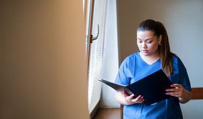 Young hispanic nurse reading a clinical report in a hospital hallway, wearing blue uniform.