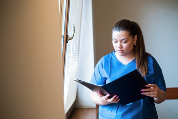 Young hispanic nurse reading a clinical report in a hospital hallway, wearing blue uniform.
