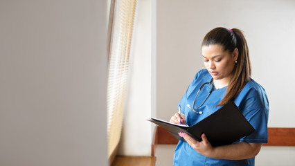 Young female hispanic doctor reading and filling a report form, standing next to a window in a hospital hallway