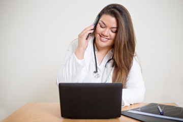 Happy smiling young female hispanic doctor sitting behind the desk and talking on the mobile phone