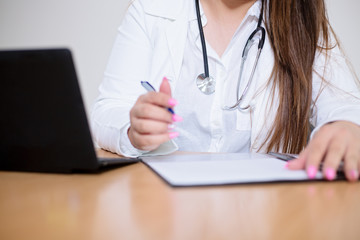 Close-up of young female doctor sitting behind the desk filling a form