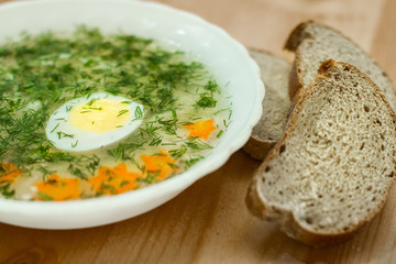 Soup with egg, vegetables, dill in white plate on a wooden table. Three slices of bread.