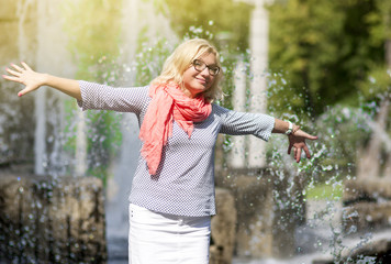 Portrait of Funny Mature Middle Aged Smiling Blond Woman Wearing Spectacles Posing Outdoors in Park. Showing Outstretched hands Against Nature Background.