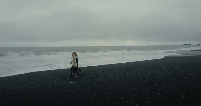 Copter flying around young couple in icelandic sweater walking on black volcanic beach in Iceland and looking on waves.