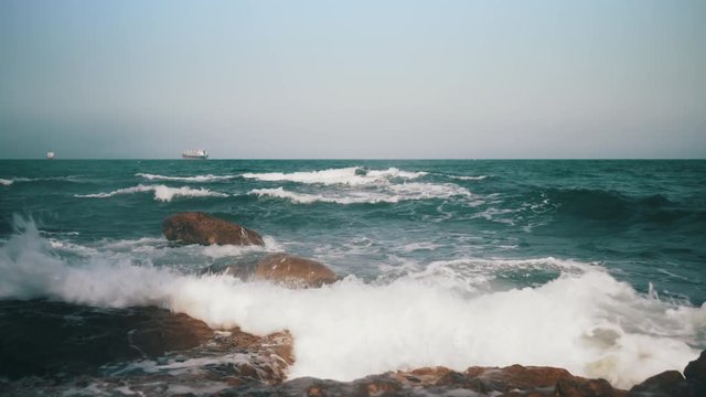 Dynamic picture. Large waves on the sea. Sandy beach with large stones on the beach. Waves break on stones. Lonely cargo ship in the distance. Ship raid