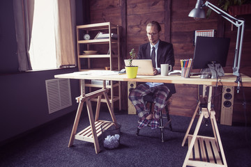 Freelancer at working place at home. Man in jacket working with laptop, his legs in pajamas under table. Toned image.
