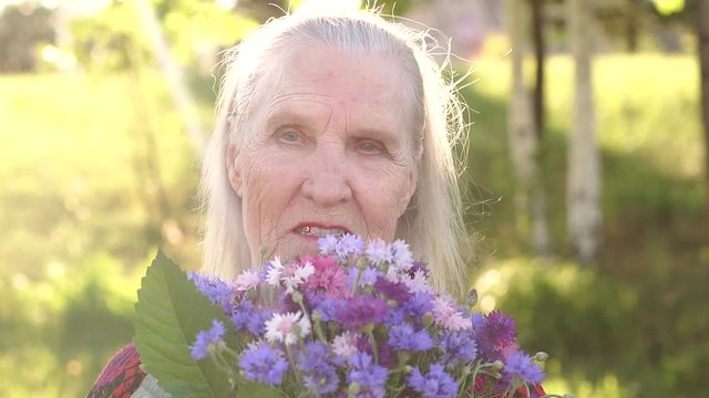 Portrait of woman with wrinkles and gray hair with a bouquet of wildflowers in a Sunny Park. Slow motion. Portrait of grandmother with a bouquet of flowers.