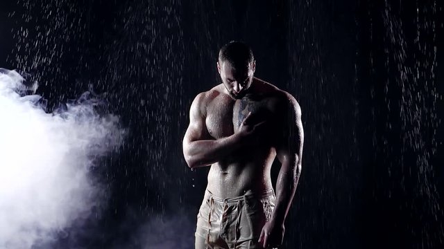 man with sexy athletic body alone in the dark stands under water jets. he caresses your arm muscles and posing. on a black background mist. in his seductive body flow streams of water