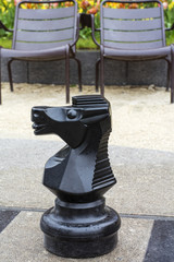 Black chess knight is on the street chessboard with two brown chairs as background © Anastasia Pestova