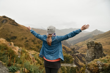 Happy young woman with arms raised out and up standing in the mountain valley in New Zealand near dangerous road. South Island. Cloud rainy day