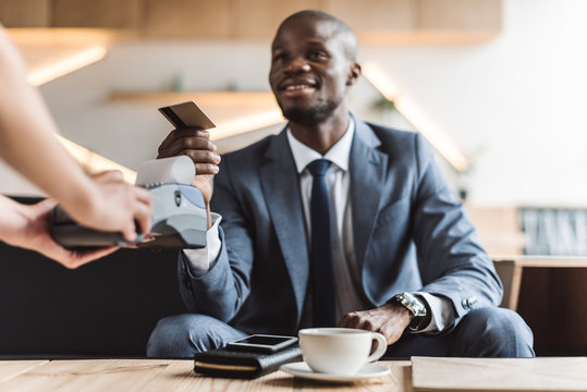 handsome smiling african american businessman paying with credit card in cafe