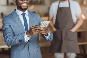 cropped view of handsome african american businessman holding and counting money, waiter standing behind in cafe
