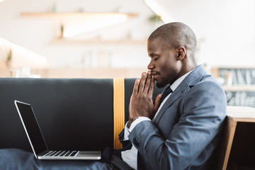 smiling african american businessman with closed eyes praying with laptop