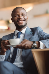 handsome smiling african american businessman sitting with cup of coffee in cafe