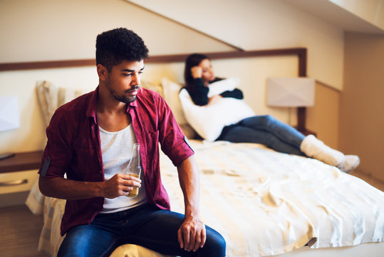 Man drinking alcohol while a sad wife is hugging a pillow in bed.