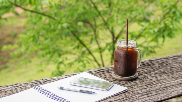 Iced Americano Coffee, Smartphone, A Pen, And A Book On A Wooden Table.