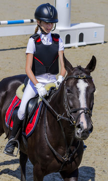 Young girl jumping with black horse. Blond pretty little girl going jump a hurdle in a competition. Girl with crow horse during equestrian showjumping. Bright sunny day.