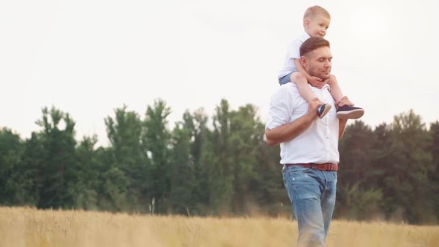 Happy father and little son walking in summer field and having fun.Son sits on his father's neck.Happy young family spending time together outside in green nature.
