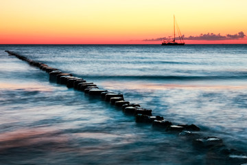 Wooden groynes and sailing boat. Red sunset and long exposure.