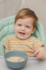 little blond boy in a striped T-shirt eats porridge with a spoon from a gray plate