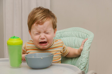 little blond boy in a striped T-shirt does not want to eat a porridge with a spoon from a gray plate.