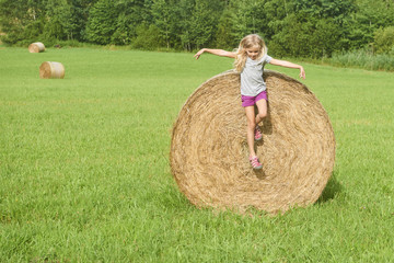 Naklejka na ściany i meble Child blond Girl by straw hay bale in field. Children summer activities 