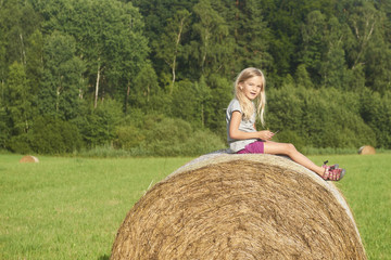 Naklejka na ściany i meble Child blond Girl by straw hay bale in field. Children summer activities 