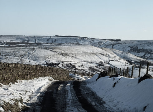 view of yorkshire moors covered in snow with a country road going downhill and the pennines on the horizon with paths and fields on a bright sunny day