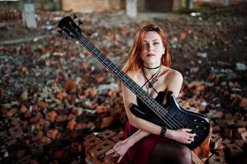 Fototapeta na wymiar Red haired punk girl wear on black and red skirt, with bass guitar at abadoned place. Portrait of gothic woman musician.