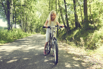 Naklejka premium Child blond girl riding a bicycle on a bike path in summer