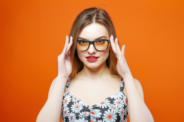 Naklejka premium Portrait of Beautiful Female Model in Eyeglasses on Orange Background. Attractive Brunette with Sensual Lips and Dress with Flowers is Posing in Studio.